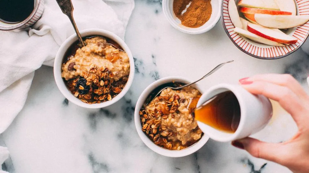 Two bowls of oatmeal with granola, sliced apples, grapes, and coffee on a marble table