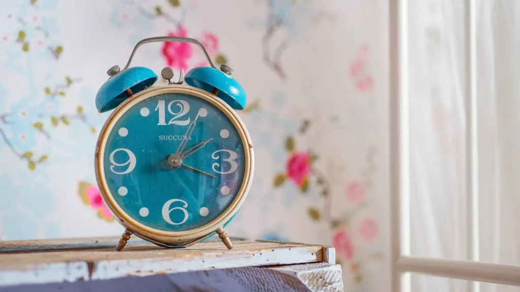 A vintage teal alarm clock on a wooden shelf against floral wallpaper